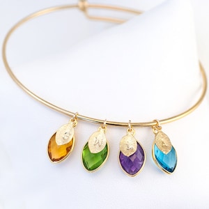 May include: Gold bangle bracelet with four charms. Each charm is a gold leaf with a letter engraved on it and a gemstone in the shape of a teardrop. The gemstones are orange, green, purple, and blue.