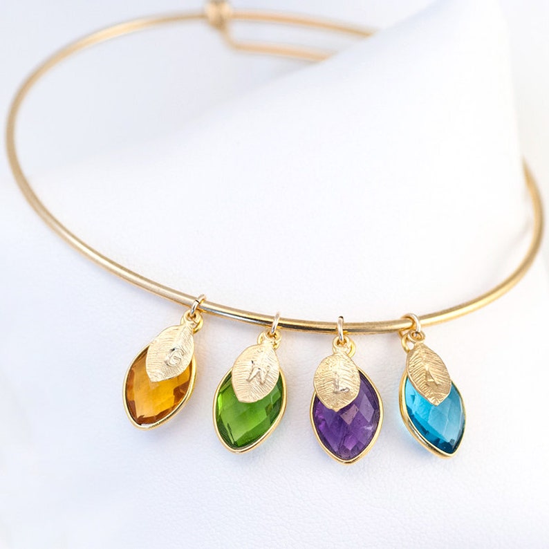 May include: Gold bangle bracelet with four charms. Each charm is a gold leaf with a letter engraved on it and a gemstone in the shape of a teardrop. The gemstones are orange, green, purple, and blue.