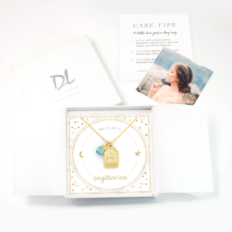 May include: A gold necklace with a Sagittarius zodiac sign charm and a turquoise gemstone. The necklace is displayed in a white box with a gold star pattern and the words "Sagittarius" and "Nov. 22 - Dec. 21".