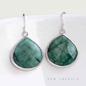May include: A pair of silver dangle earrings with teardrop-shaped raw emerald gemstones. The earrings have a simple design and are perfect for everyday wear.