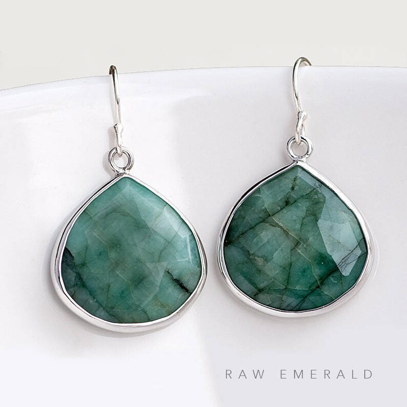 May include: A pair of silver dangle earrings with teardrop-shaped raw emerald gemstones. The earrings have a simple design and are perfect for everyday wear.