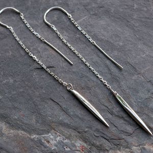 Op de afbeelding: Een paar zilveren oorhangers met een lange, dunne, puntige druppel. De oorhangers zijn gemaakt van een delicate ketting en hebben een eenvoudig, minimalistisch ontwerp.