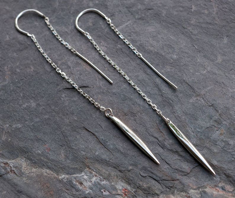 Op de afbeelding: Een paar zilveren oorhangers met een lange, dunne, puntige druppel. De oorhangers zijn gemaakt van een delicate ketting en hebben een eenvoudig, minimalistisch ontwerp.