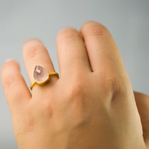 K&ouml;nnte beinhalten: Ein goldfarbener Ring mit einem tropfenf&ouml;rmigen, hellrosa Edelstein. Der Ring hat ein schlichtes, elegantes Design, das den facettierten Stein hervorhebt. Das Band ist d&uuml;nn und goldfarben. Der Ring wird am Finger getragen.