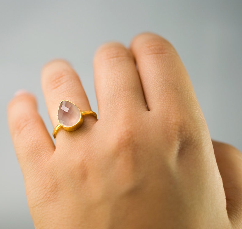 K&ouml;nnte beinhalten: Ein goldfarbener Ring mit einem tropfenf&ouml;rmigen, hellrosa Edelstein. Der Ring hat ein schlichtes, elegantes Design, das den facettierten Stein hervorhebt. Das Band ist d&uuml;nn und goldfarben. Der Ring wird am Finger getragen.
