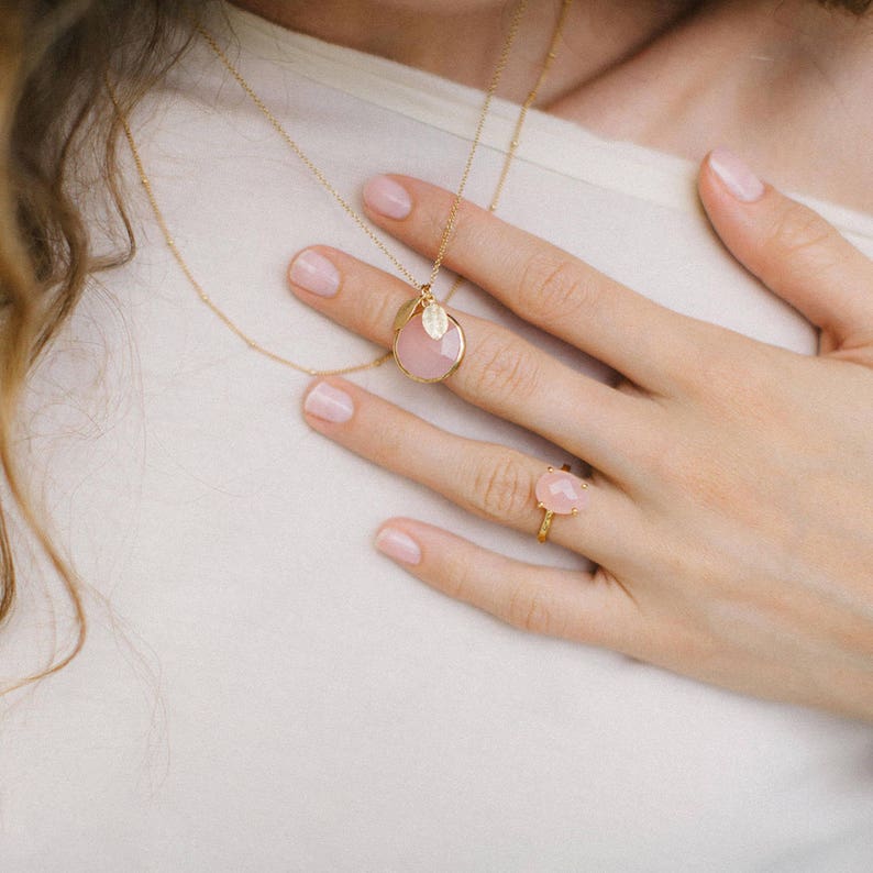 May include: A gold necklace with a pink stone pendant and a small leaf charm. A matching pink stone ring is on the finger of a hand.