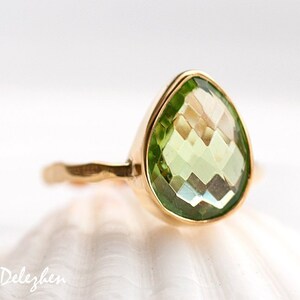 May include: A gold ring with a pear-shaped green gemstone. The ring is set on a white seashell.