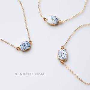 May include: Three gold necklaces with white dendrite opal stones. The necklaces are on a white background. The text "DENDRITE OPAL" is written below the necklaces.