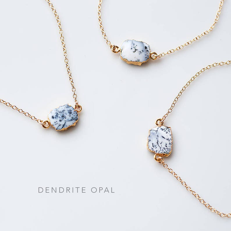 May include: Three gold necklaces with white dendrite opal stones. The necklaces are on a white background. The text "DENDRITE OPAL" is written below the necklaces.