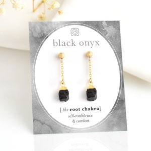 May include: Black onyx earrings with gold chains and beads. The earrings are on a white card with the text "black onyx" and "[the root chakra] self-confidence & comfort".