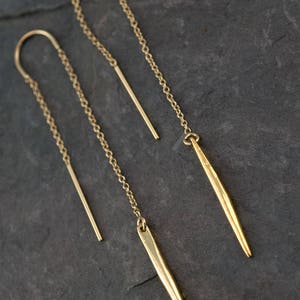Op de afbeelding: Een paar gouden oorbellen met een lange ketting en een puntige, geometrische hanger. De oorbellen zijn gemaakt van metaal en hebben een minimalistisch ontwerp.