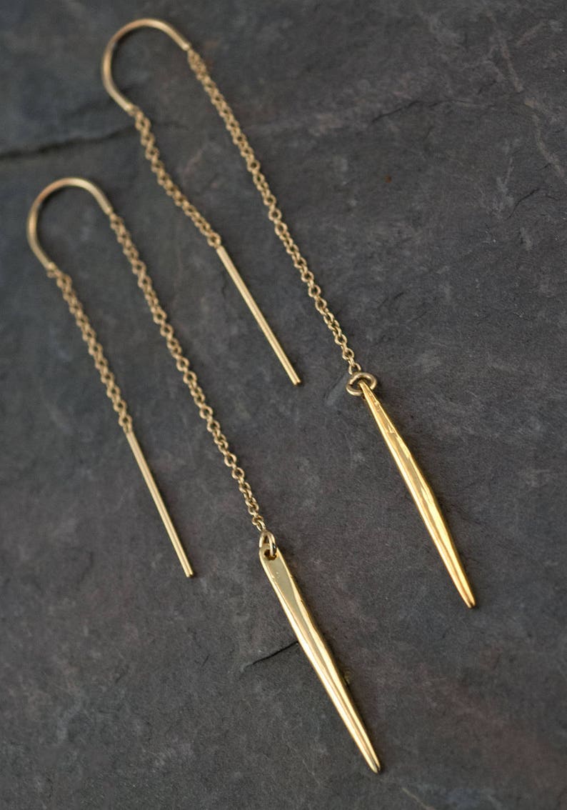 Op de afbeelding: Een paar gouden oorbellen met een lange ketting en een puntige, geometrische hanger. De oorbellen zijn gemaakt van metaal en hebben een minimalistisch ontwerp.