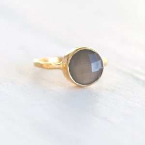 May include: A gold ring with a gray gemstone. The ring has a textured band and a round, faceted stone.