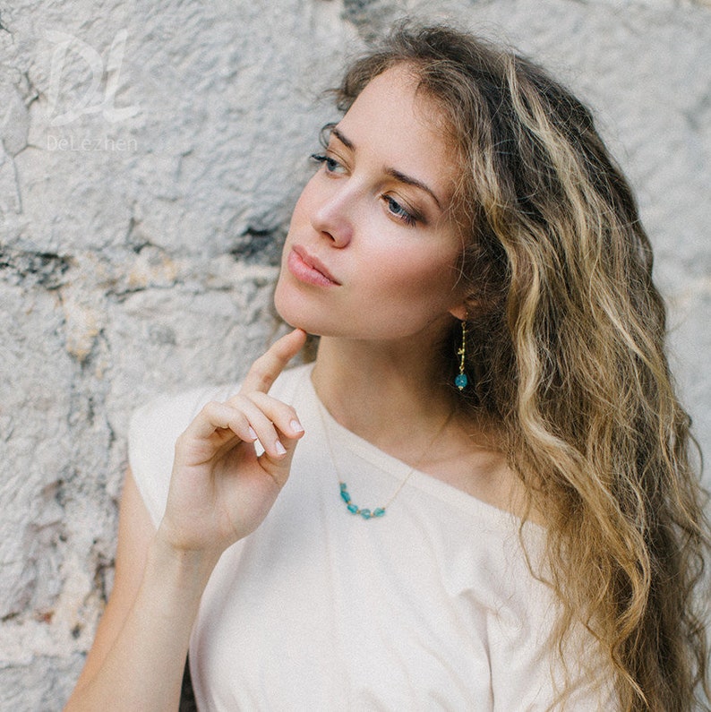 May include: A woman with long, wavy blonde hair wearing a white shirt and a gold necklace with turquoise stones. She is also wearing turquoise earrings.