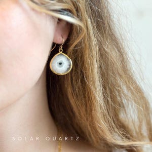 May include: Gold earrings with a white and gray solar quartz stone. The stone has a natural, crystal-like appearance.  The earrings are a teardrop shape and have a simple, elegant design.  "SOLAR QUARTZ" is written in the bottom of the image.