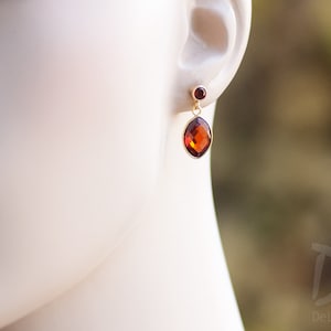 May include: Gold drop earrings with a large, faceted, oval amber gemstone and a smaller, round red gemstone. The earrings are displayed on a mannequin's ear, highlighting their design and colour. The earrings are a classic jewellery item.