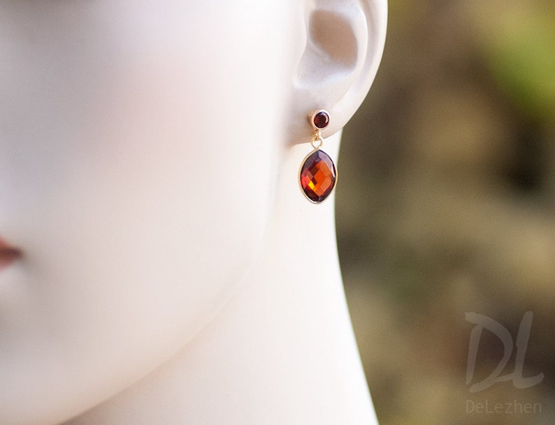 May include: Gold drop earrings with a large, faceted, oval amber gemstone and a smaller, round red gemstone. The earrings are displayed on a mannequin's ear, highlighting their design and colour. The earrings are a classic jewellery item.