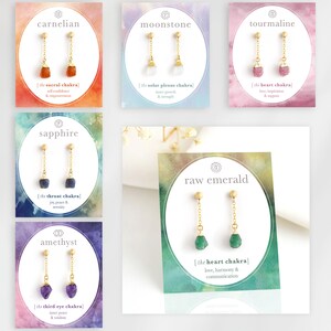 以下が含まれることがあります： 水彩画の背景が付いたカードに、10組の宝石のイヤリングが飾られています。各カードには、宝石の名前、対応するチャクラ、チャクラの意味についての簡単な説明が記載されています。宝石には、カーネリアン、ムーンストーン、トルマリン、サファイア、原石のエメラルド、アメジストが含まれています。