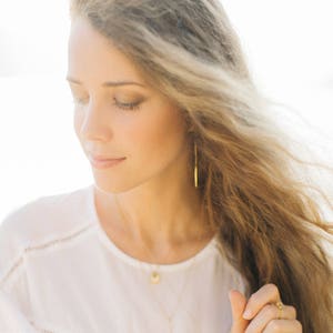 Op de afbeelding: Een vrouw met lang, golvend blond haar draagt een wit shirt en gouden sieraden. Ze draagt een ketting met een klein hangertje en lange, dunne gouden oorbellen.