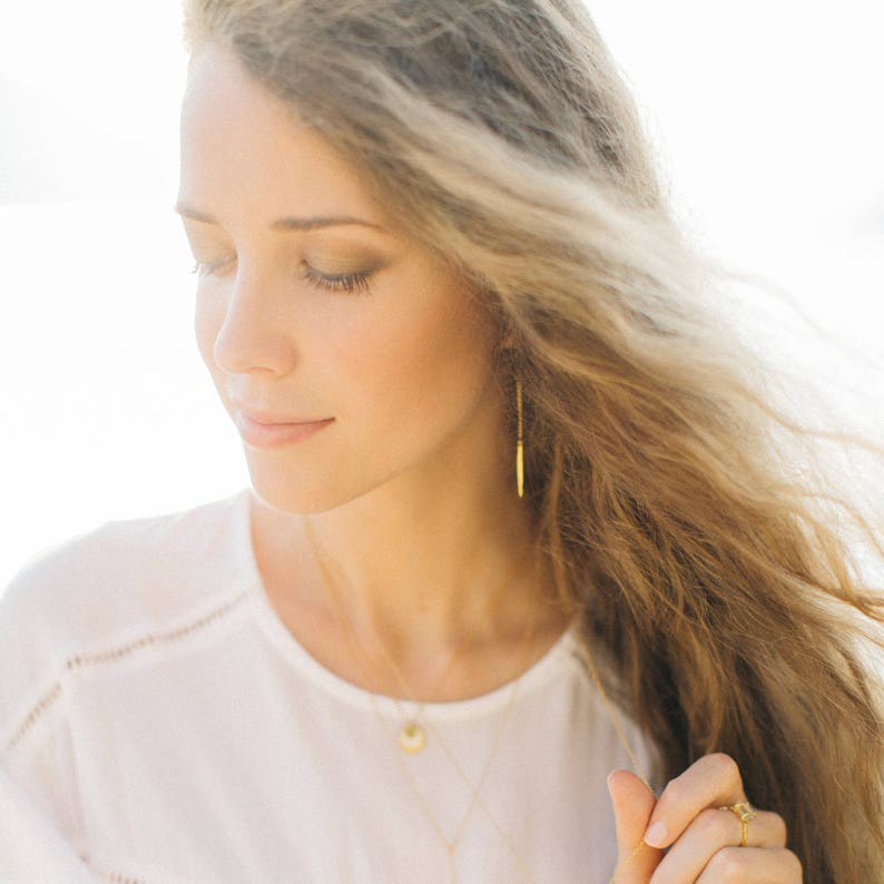 Op de afbeelding: Een vrouw met lang, golvend blond haar draagt een wit shirt en gouden sieraden. Ze draagt een ketting met een klein hangertje en lange, dunne gouden oorbellen.