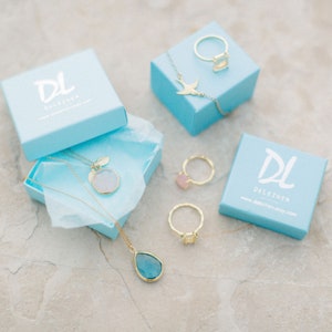 May include: Two light blue boxes with the logo "DL DELEZHEN" and the website address "www.delezhen.etsy.com". The boxes contain gold necklaces with a round and teardrop shaped pendant, a gold ring with a square shaped stone, and a gold ring with a round shaped stone.