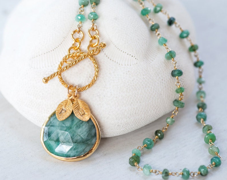 May include: A gold chain necklace with a large emerald green gemstone pendant. The pendant is surrounded by gold leaf accents. The chain is made of small emerald green beads.