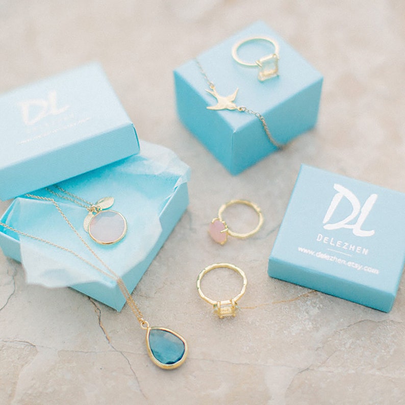 Pu&ograve; includere: Un set di gioielli delicati in oro con collane e anelli con vari accenti di pietre preziose. I gioielli sono esposti in scatole blu chiaro con il logo "DL DELEZHEN" su una delle scatole.