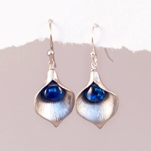 Puede incluir: Un par de pendientes de plata con una piedra preciosa azul en el centro de cada pendiente. Los pendientes tienen forma de p&eacute;talo de flor.