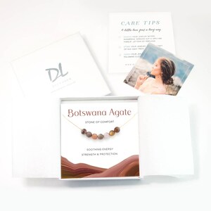 Puede incluir: Una caja blanca con un collar de oro en su interior. El collar tiene un dije de &aacute;gata marr&oacute;n con las palabras "Botswana Agate" y "Stone of Comfort" impresas en la caja. La caja tambi&eacute;n tiene las palabras "Soothing Energy, Strength & Protection" impresas en ella.