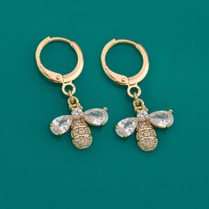 Puede incluir: Pendientes de aro dorados con un dije de abeja colgante. La abeja está adornada con piedras de circonita cúbica transparentes.