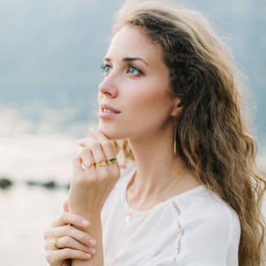 Op de afbeelding: Een vrouw met lang, golvend bruin haar draagt een wit shirt en gouden sieraden. Ze draagt een gouden ketting met een klein hangertje, een paar gouden oorbellen en drie gouden ringen met gekleurde stenen.