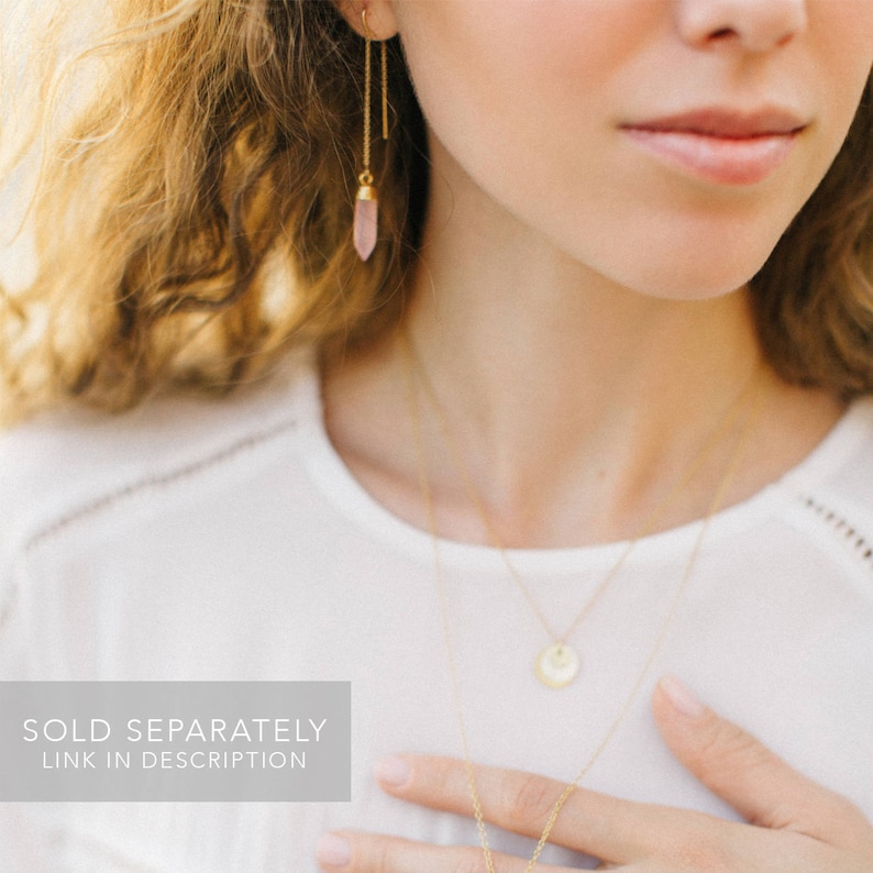 May include: A woman wearing a white shirt with a gold chain necklace and a pair of gold earrings with pink crystal accents. The text "SOLD SEPARATELY LINK IN DESCRIPTION" is at the bottom of the image.