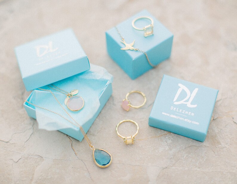 May include: Four light blue boxes with gold jewelry inside. The boxes have the logo "DL DELEZHEN" printed on them. The jewelry includes a necklace with a round pendant, a ring with a square stone, and a ring with a round stone.