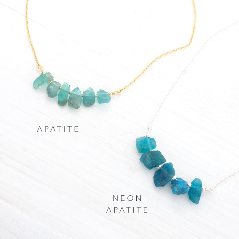 May include: Two necklaces with blue apatite stones. The top necklace has a gold chain and the bottom necklace has a silver chain. The top necklace is labeled "Apatite" and the bottom necklace is labeled "Neon Apatite".