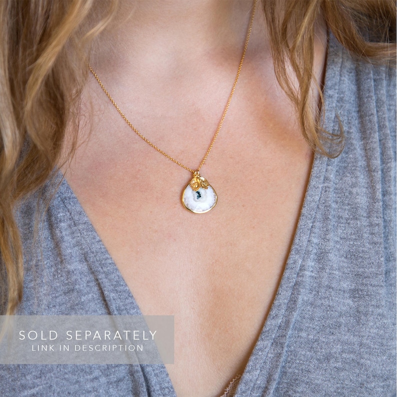 May include: A gold chain necklace with a white porcelain pendant featuring a small blue gemstone and gold leaf accents. The pendant is shaped like a teardrop.