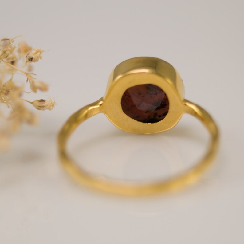 May include: A gold ring with a round, dark red gemstone set in a bezel setting. The ring is simple and elegant.