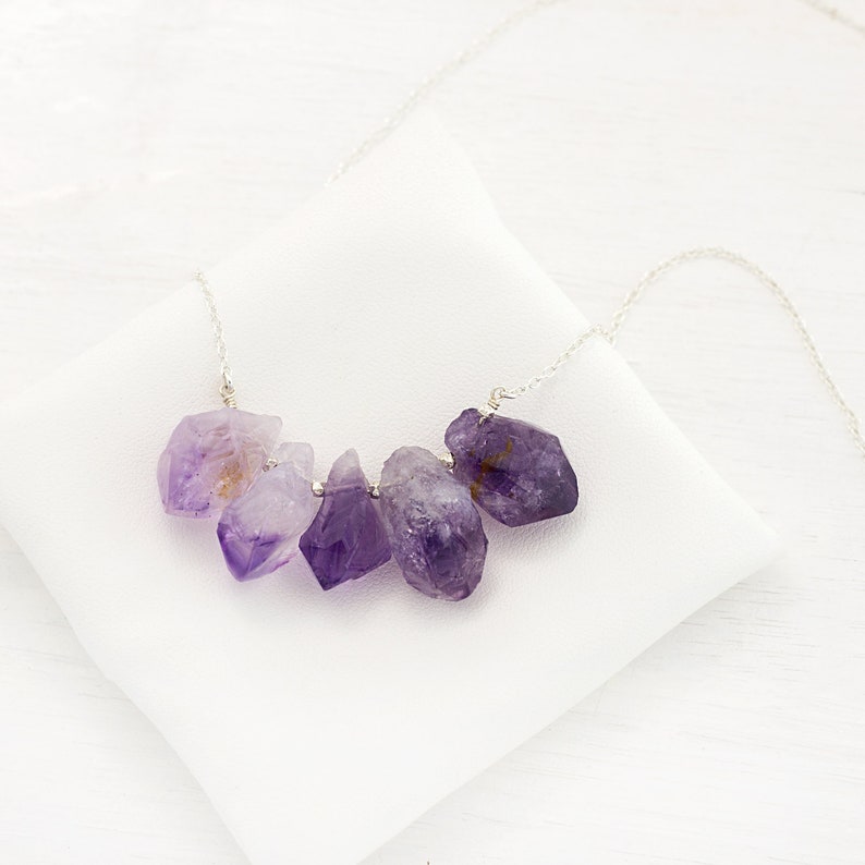 May include: A silver chain necklace with a cluster of raw amethyst crystals in shades of purple. The crystals are irregular in shape and have a natural, rough texture.
