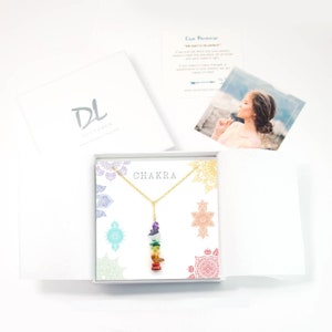 May include: A gold chain necklace with a seven-stone chakra pendant. The stones are stacked in a rainbow order, starting with red at the bottom and ending with purple at the top. The necklace is displayed in a white box with a mandala pattern and the word "CHAKRA" printed on the front.
