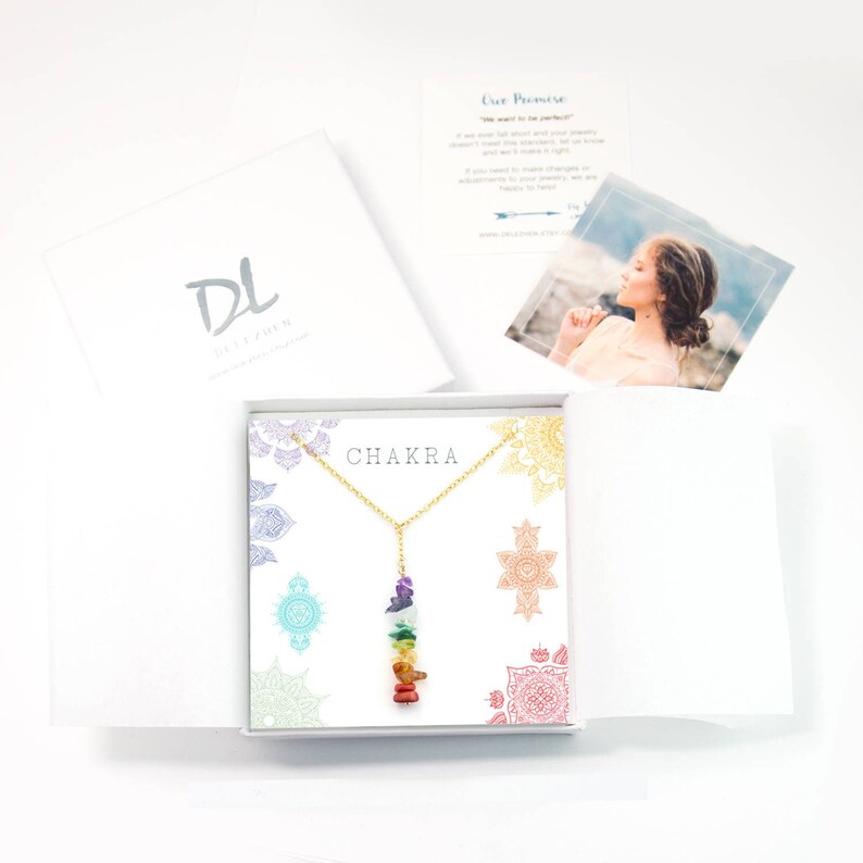 May include: A gold chain necklace with a seven-stone chakra pendant. The stones are stacked in a rainbow order, starting with red at the bottom and ending with purple at the top. The necklace is displayed in a white box with a mandala pattern and the word "CHAKRA" printed on the front.