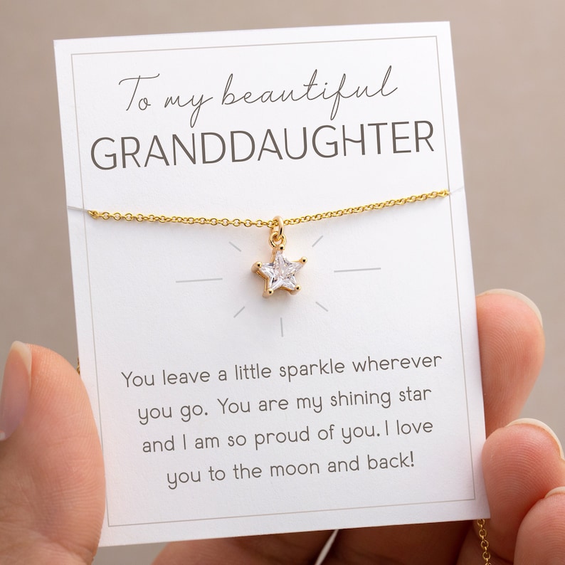 Puede incluir: Un collar dorado con un colgante en forma de estrella, presentado en una tarjeta blanca. La tarjeta dice "To my beautiful GRANDDAUGHTER" e incluye un mensaje afectuoso. El collar es sostenido por una persona.