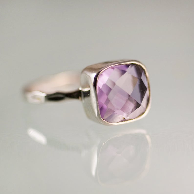May include: A silver ring with a square-shaped amethyst gemstone. The gemstone is a light purple color and has a faceted cut.