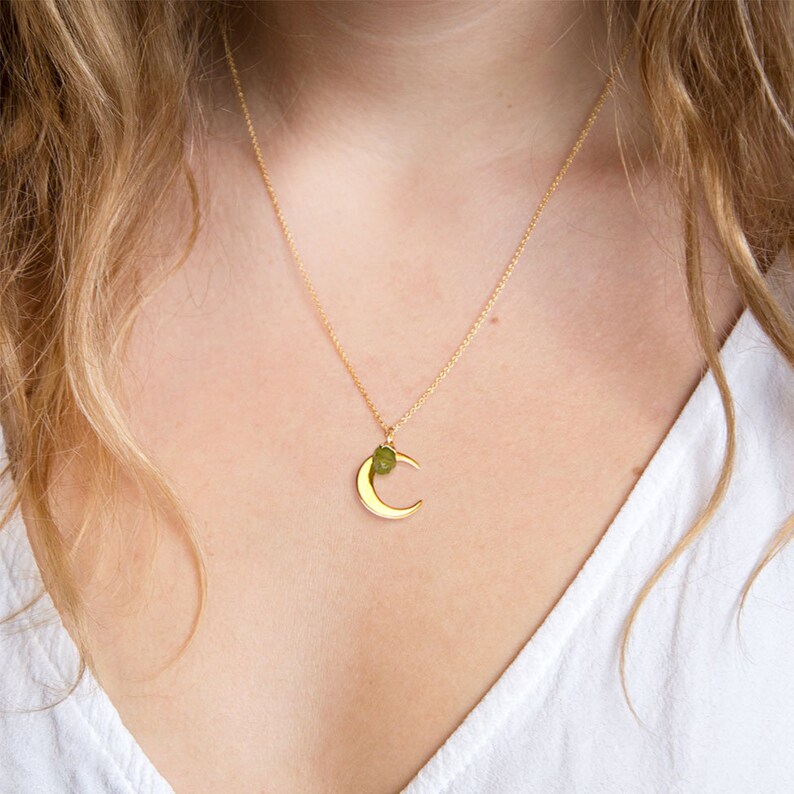May include: A gold crescent moon necklace with a small green gemstone accent. The necklace is worn by a person with long blonde hair.