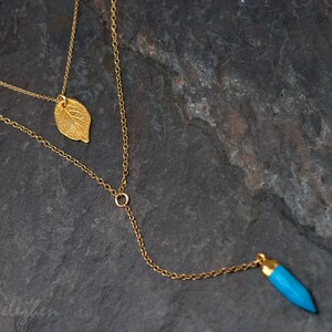 May include: A gold chain necklace with two pendants. One pendant is a gold leaf and the other is a blue teardrop-shaped stone.