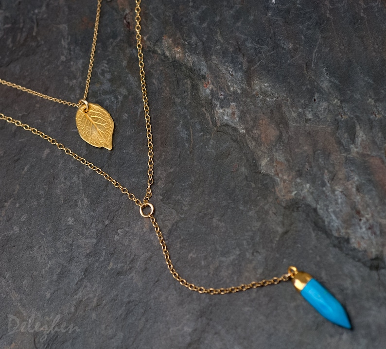 May include: A gold chain necklace with two pendants. One pendant is a gold leaf and the other is a blue teardrop-shaped stone.