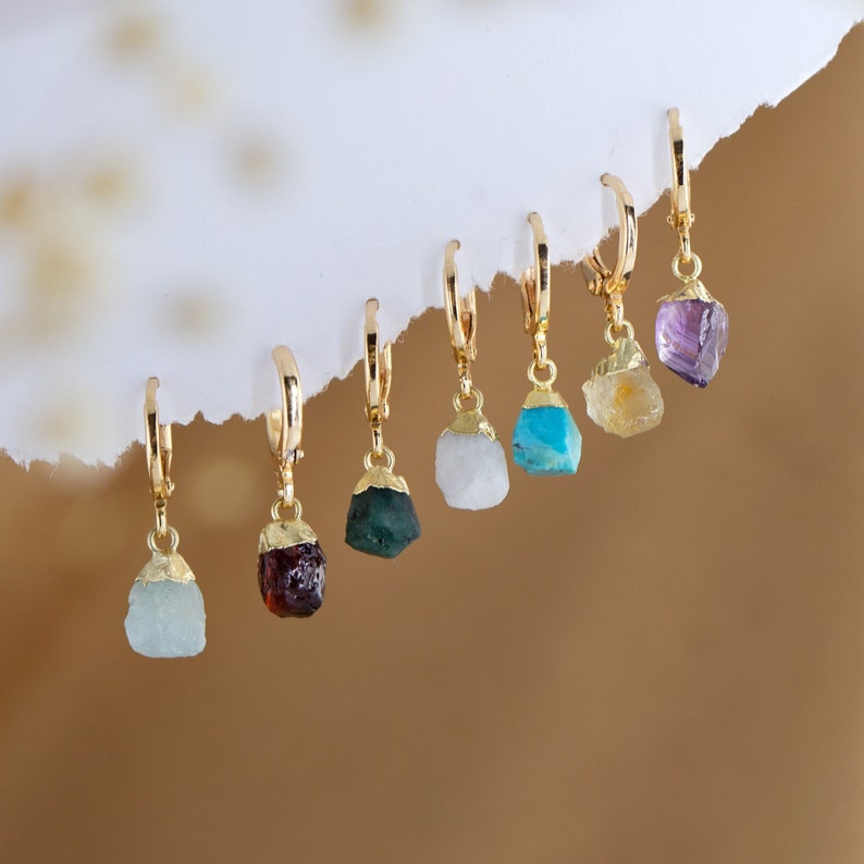 May include: A set of seven gold hoop earrings with different colored gemstone charms. The charms are in the shape of rough, natural stones. The colors of the stones are purple, yellow, blue, white, green, and red.