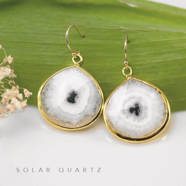 May include: A pair of gold earrings with white and black solar quartz stones. The stones are round and have a teardrop shape. The earrings are hanging from gold hooks.