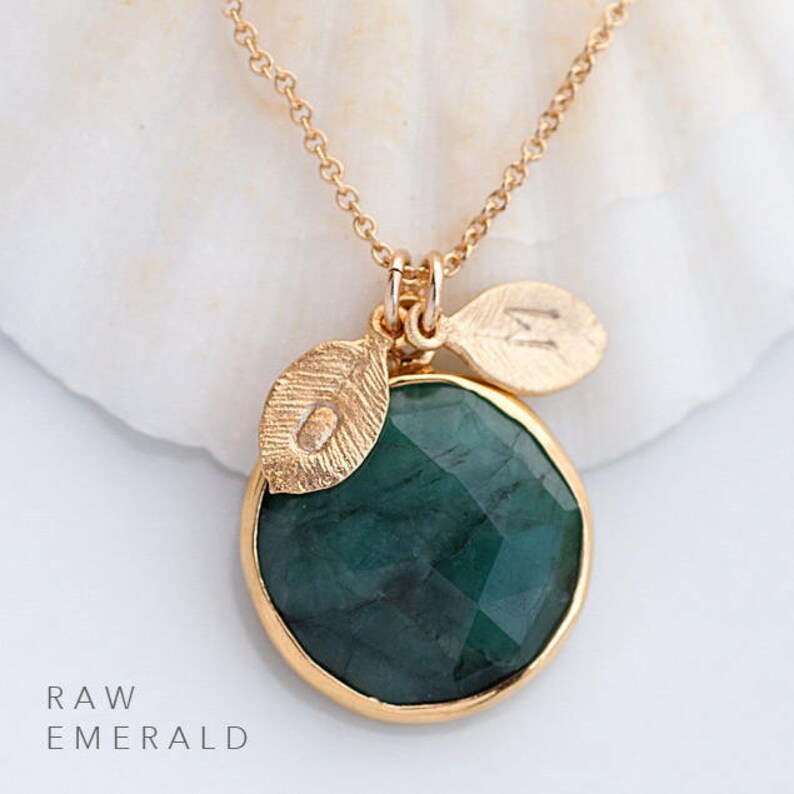 May include: A gold necklace with a raw emerald pendant and two small gold leaf charms. The pendant is round and has a faceted surface. The necklace is on a white background. RAW EMERALD is written below the pendant.