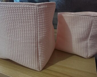 Può includere: Due cesti portaoggetti in tessuto rosa chiaro a trama waffle di diverse dimensioni sono appoggiati su una superficie di legno. Il cesto più grande è rettangolare, mentre quello più piccolo è quadrato. I cesti hanno un aspetto strutturato.