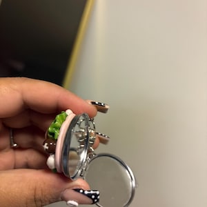 K&ouml;nnte beinhalten: Ein silberfarbener, aufklappbarer Kompaktspiegel mit zwei kreisf&ouml;rmigen Spiegeln. Die Oberseite des Kompakts weist ein Miniatur-Design mit bunten Lebensmitteln auf, darunter gr&uuml;ner Salat, braune Fleischb&auml;llchen und rosa Elemente. Der Kompaktspiegel ist ge&ouml;ffnet und zeigt die Spiegelfl&auml;chen.