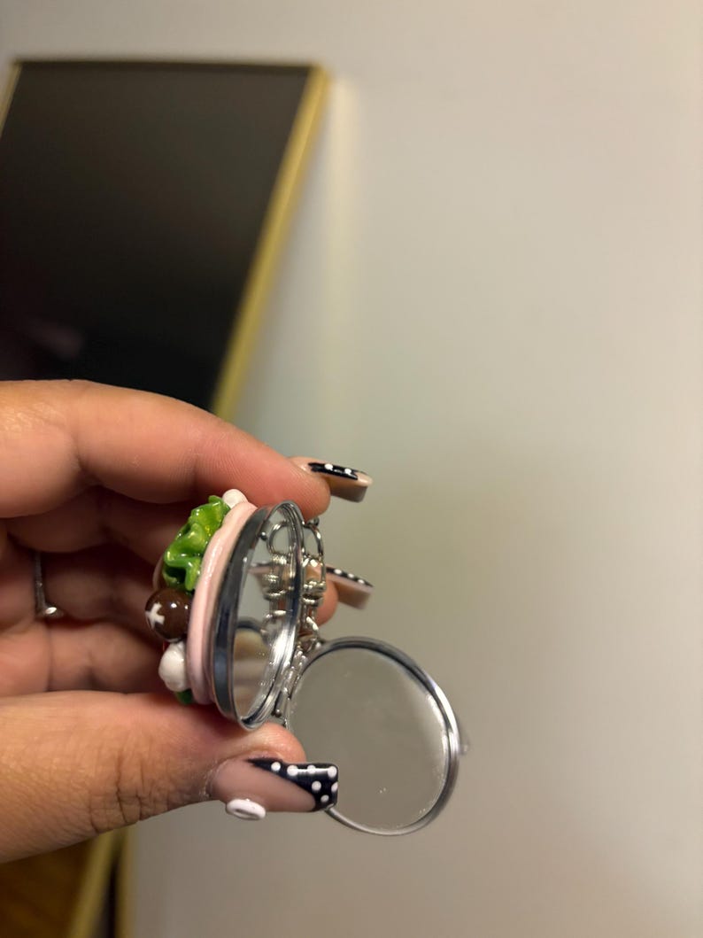 K&ouml;nnte beinhalten: Ein silberfarbener, aufklappbarer Kompaktspiegel mit zwei kreisf&ouml;rmigen Spiegeln. Die Oberseite des Kompakts weist ein Miniatur-Design mit bunten Lebensmitteln auf, darunter gr&uuml;ner Salat, braune Fleischb&auml;llchen und rosa Elemente. Der Kompaktspiegel ist ge&ouml;ffnet und zeigt die Spiegelfl&auml;chen.